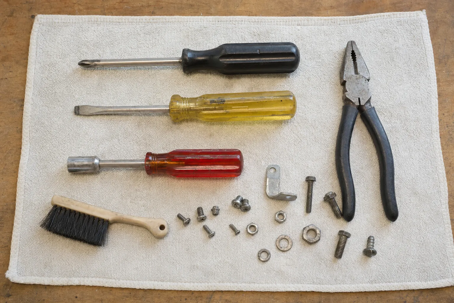 Two screwdrivers and a nut driver in a row above an L-bracket, loose screws, washers and nuts, with pliers and a hand brush