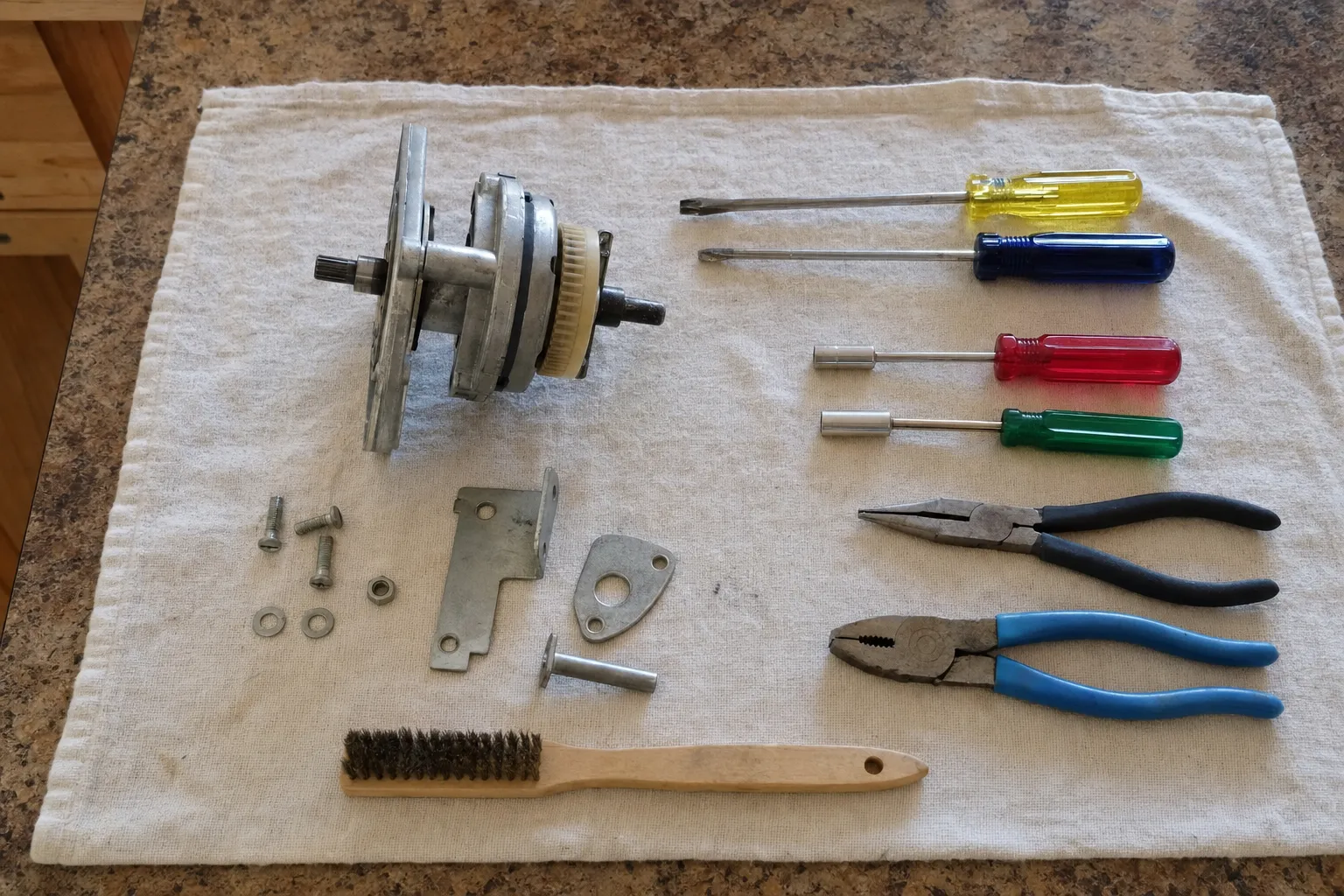 A small geared motor assembly on a counter towel beside two nut drivers, two screwdrivers, two pairs of pliers and a wire brush