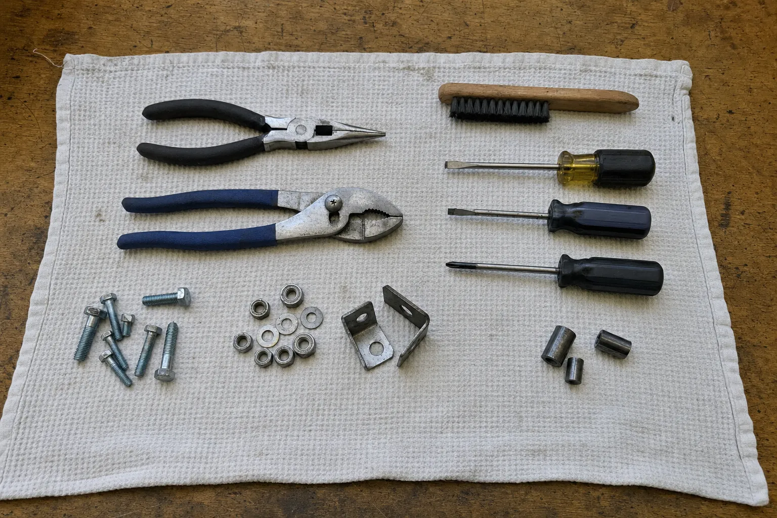Needle-nose and slip-joint pliers, three screwdrivers and a wire brush above hex bolts, nuts, two angle brackets and three bushings